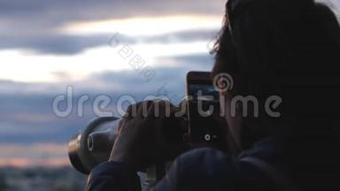 旅行者年轻的女游客在日落时用投币式望远镜观察城市，并用手机拍照